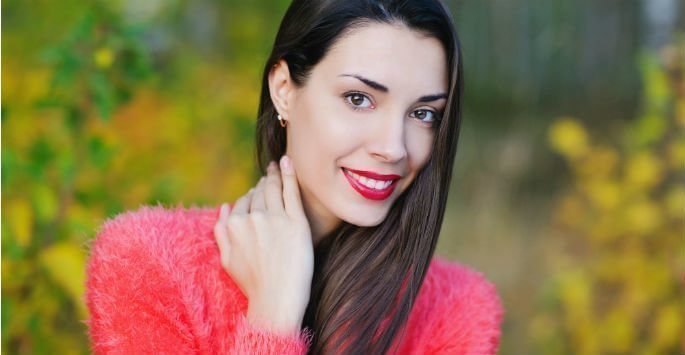 Woman with long brown hair outdoors smiling in a pink sweater