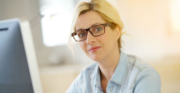 Mature woman with stringy blonde hair wearing glasses sitting at a computer