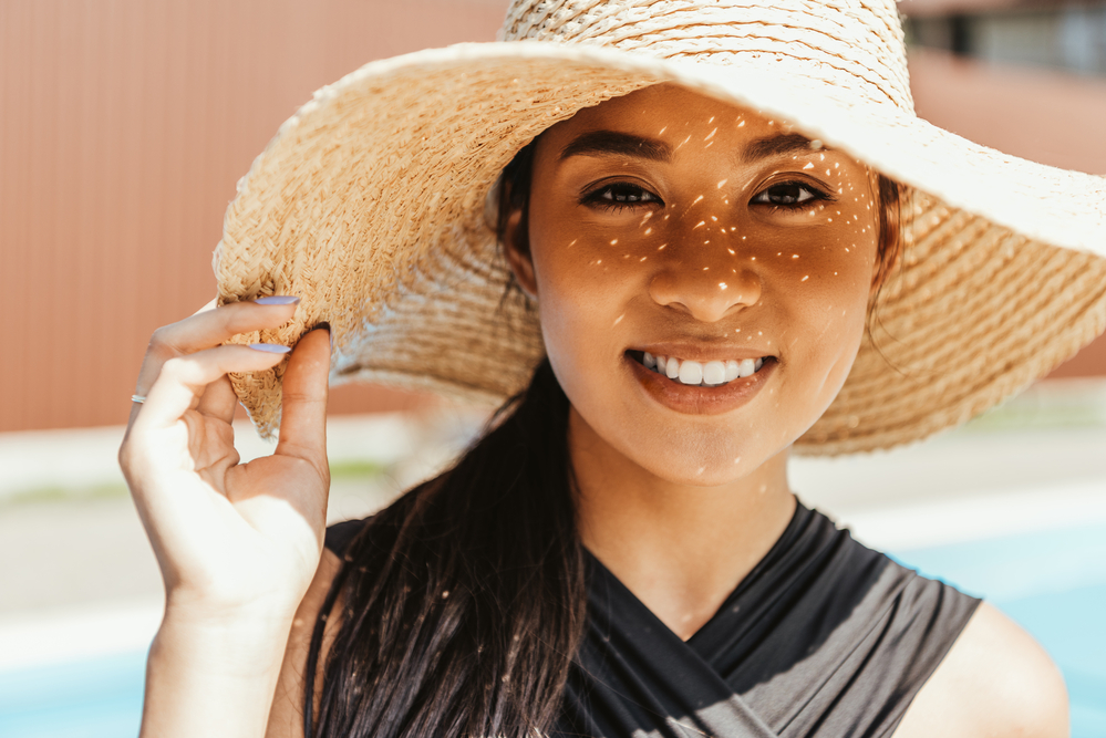 Smiling woman outdoors wearing a wide-brimmed hat with sun shining through