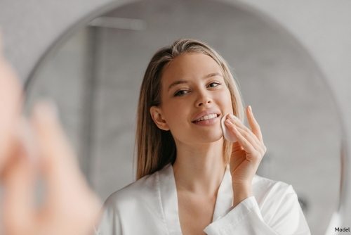 Woman in a robe using skincare products on her skin while looking in the mirror and smiling