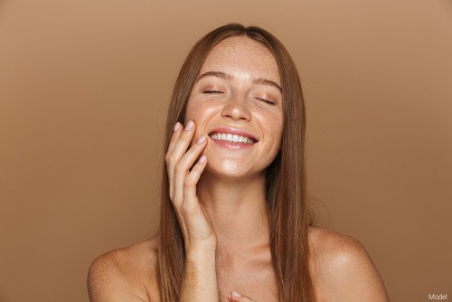 Joyful young woman with long, straight brown hair smiling and gently touching her cheek